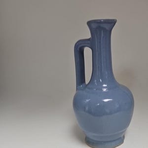 May include: A blue ceramic vase with a handle. The vase has a rounded body, a narrow neck, and a small opening. The handle is attached to the neck and the body. The vase is approximately 8 inches tall.