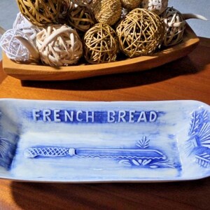 May include: An elongated white ceramic serving tray with blue painted accents. The tray features the embossed text FRENCH BREAD and a relief image of a serrated bread knife in the center, with decorative wheat motifs on both ends.