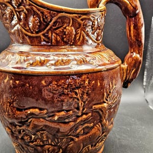 May include: Brown ceramic pitcher with a detailed relief design. The pitcher features a curved handle and a spout. The relief depicts a landscape with trees, foliage, and grapevines. A decorative piece for serving beverages.