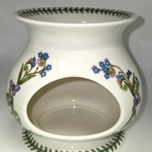 May include: A white ceramic oil warmer with a wide, rounded body and a large circular opening in the front. The exterior features a botanical pattern of blue flowers, green leaves, and small purple butterflies, with a green leaf border at the rim and base.