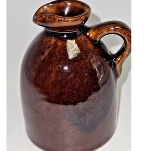 May include: A small, brown ceramic jug with a handle and a spout. The jug has a glossy finish and a rounded body. The handle is arched, and the spout is small and curved. The jug appears to be vintage or antique.