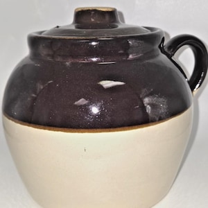 May include: A round ceramic crock with a lid and a single side handle. The upper portion of the vessel is finished in a dark brown glaze, while the lower portion features a smooth, light cream-colored base. The lid has a small central knob.