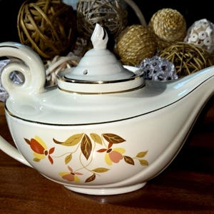 May include: A cream-colored ceramic teapot featuring a gold-trimmed lid and a curved handle. The body is decorated with a floral pattern in shades of orange, yellow, and brown. The lid has a pointed finial and a small steam vent hole.