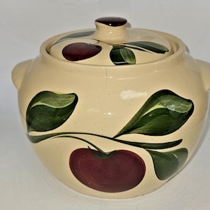 May include: Cream-colored ceramic lidded pot with a hand-painted design of dark red cherries and green leaves. The pot has two side handles and a small knob on the lid. A charming piece for kitchen decor.