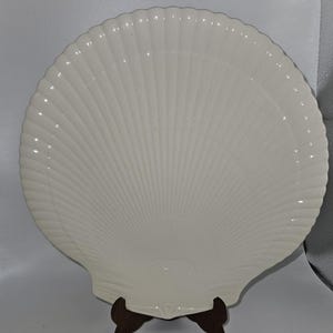 May include: A large, off-white seashell-shaped serving platter. The platter has a ribbed, fan-like design radiating from the center. It is displayed on a dark brown wooden stand, showcasing its elegant form. A decorative piece for coastal decor.