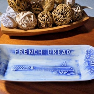 May include: A rectangular white ceramic serving tray with blue painted accents. The center features the embossed text FRENCH BREAD and a relief image of a serrated bread knife. The ends are decorated with embossed wheat stalks.