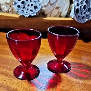 May include: Two ruby red stemmed glasses with a rounded bowl and a circular base. The glasses are made of transparent glass and sit on a wooden surface. Decorative elements are in the background.