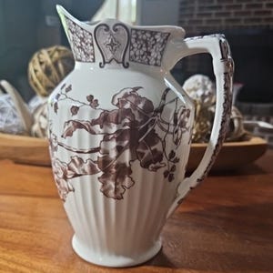 May include: A white ceramic pitcher with a brown floral design. The pitcher has a curved spout, a ribbed body, and a decorative handle. The floral pattern includes leaves and flowers. The pitcher is likely used for serving beverages.
