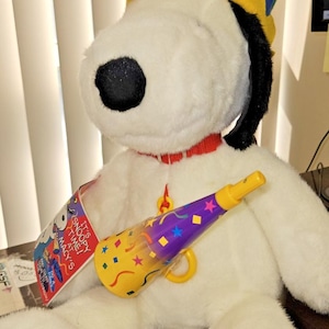May include: A white plush Snoopy dog wearing a colorful jester hat with yellow, blue, green, and red sections. The plush has a red collar and holds a purple and yellow party horn with confetti patterns. A tag reads IT'S SNOOPY TIME! AT MACY'S.
