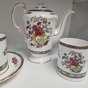 May include: A white porcelain tea set with floral designs. The set includes a teapot, a teacup, and a saucer. The teapot has a curved handle and spout with gold trim. The teacup and saucer also have gold trim. The floral design includes red, purple, and yellow flowers.