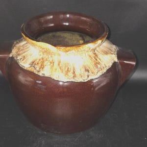 May include: A dark brown ceramic pot with two side handles and a wide, circular opening. The upper portion of the pot features a textured, cream-colored glaze that transitions into the smooth, glossy dark brown finish of the main body.