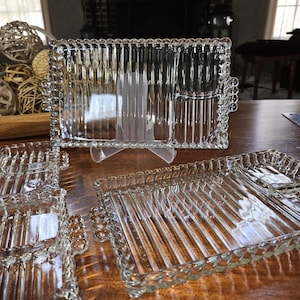 May include: A set of clear glass rectangular serving trays featuring a vertical ribbed pattern on the base and a decorative beaded edge. Each tray includes a small partitioned section on one side and integrated handles with a matching beaded design.