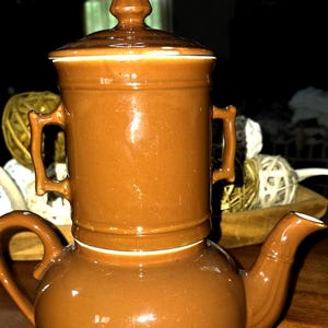 May include: A brown ceramic teapot with a stacked, two-part design. The base is rounded with a curved handle and spout, while the upper section is cylindrical with two side handles and a matching brown lid featuring a pointed finial top.