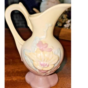 May include: A small, cream-colored ceramic pitcher with a curved handle and spout. The pitcher features a raised floral design in shades of pink, yellow, and blue. The base is a soft pink color. A decorative piece for the home.