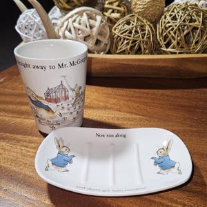 May include: A white ceramic cup and soap dish set featuring illustrations of a rabbit in a blue jacket. The cup has the text "straight away to Mr. McGregor". The soap dish has the text "Now run along and don't get into mischief".
