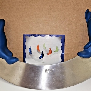 May include: A stainless steel mezzaluna knife featuring two matte blue, abstract, seated figures as handles on each end. The blade is curved and metallic, with a small logo engraved on the surface. A square card shows illustrations of similar figures.