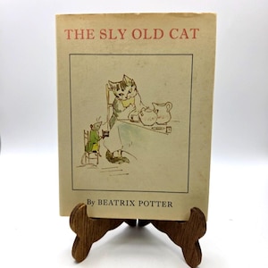 May include: A hardcover book titled The Sly Old Cat by Beatrix Potter. The cream-colored cover features a line drawing of a cat and a mouse sitting at a table with a teapot and pitcher. The title is printed in red text at the top of the book cover.
