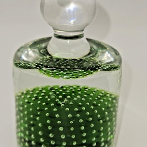 May include: A clear glass bottle with a round stopper. The bottle's base is filled with a textured, green substance dotted with small, white bubbles. The stopper and the rim of the bottle have a green tint.