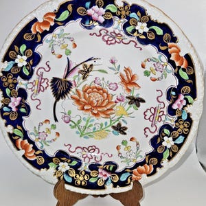 May include: A round ceramic plate featuring a white center with a large orange flower, a dark blue border, and gold accents. The design includes colorful floral motifs, green leaves, and stylized bird imagery painted across the surface.