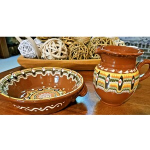 May include: A brown ceramic bowl with two handles and a matching pitcher featuring decorative bands of green, yellow, and white paint with small black and orange accents. The bowl has a central floral pattern and a wavy white line border.