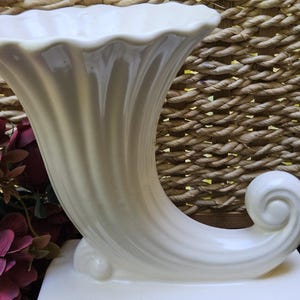 May include: Cream-colored ceramic cornucopia vase with a flared opening and a curled end. The vase sits on a rectangular base and features a ridged design on the body. The background is a woven texture.