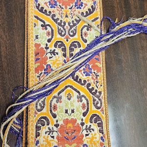 May include: A rectangular textile with a vibrant floral pattern in orange, yellow, blue, and brown. The edges are trimmed with a yellow border. Strands of blue and beige thread extend from the top.