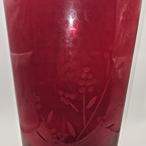 May include: A tall, ruby red glass vase with a textured surface and etched floral design. The vase has a slightly flared shape and a clear, rectangular base. The etched design features delicate flowers and leaves.