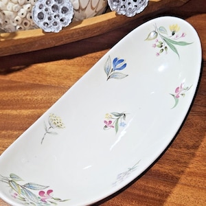 May include: An oval, off-white ceramic serving dish with a floral pattern. The dish features delicate hand-painted flowers in shades of blue, pink, yellow, and green. The dish is set on a wooden surface.