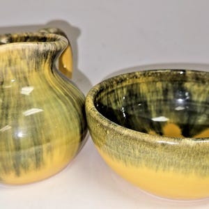 May include: A ceramic pitcher and bowl set. The pitcher has a curved handle and a spout. The bowl is round. Both pieces are glazed with a yellow, green, and black pattern. The set is likely for serving.