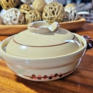 May include: A round, beige ceramic casserole dish with a matching lid. The dish features two dark brown side handles and a decorative band of small, alternating brown and pink circular patterns around the base. The lid has a central knob.