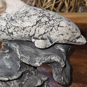 May include: A decorative figurine of a dolphin with a white and black crackled finish, positioned atop a dark gray, textured rock-like base. The dolphin is depicted in a horizontal swimming pose, showcasing a detailed, weathered surface texture.