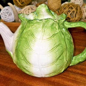 May include: A ceramic teapot shaped like a head of cabbage with a glossy finish. The body features layered green and white leaf textures, a curved handle, and a spout that resembles a pale stalk. A matching lid sits on top of the round vessel.