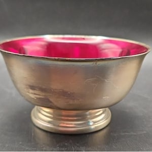 May include: A silver-toned bowl with a vibrant pink interior. The bowl has a rounded shape and sits on a small, circular pedestal base. The bowl's exterior shows some wear, adding to its vintage appeal.