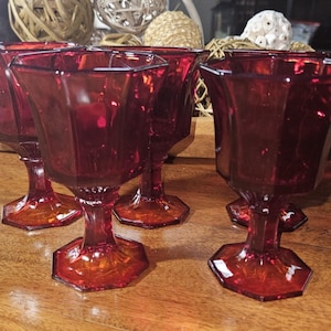 May include: A set of six ruby red vintage goblets. Each glass features a hexagonal base and a flared, faceted bowl. The stem and base are a darker shade of red. The glasses are arranged on a wooden surface.
