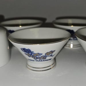 May include: A set of white ceramic sake cups and a carafe. The cups have a gold rim and a blue floral design with gold accents. The carafe and cups have Japanese text. The set is designed for serving sake.