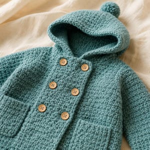 May include: A teal blue crochet hooded jacket featuring a double-breasted front closure with six light brown wooden buttons. The garment has a textured knit pattern, a hood topped with a small pom-pom, and two square patch pockets on the front.
