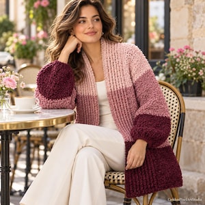 Könnte beinhalten: Eine farblich abgesetzte Strickjacke mit offener Vorderseite und gerippter Textur in den Farben Hellrosa, Altrosa und Dunkelburgund. Das Kleidungsstück hat lange, voluminöse Ärmel und einen geraden Saum, getragen über einem weißen Oberteil.