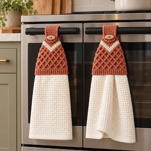 May include: Two hanging kitchen towels featuring a cream-coloured waffle weave bottom section and a rust-coloured crocheted top with a diamond pattern. Each towel is secured with a light wood button at the top, allowing it to hang from a horizontal bar.