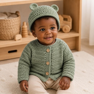 May include: A sage green crocheted baby cardigan with long sleeves and three light brown wooden buttons down the front, paired with a matching crocheted beanie hat featuring two small rounded ears on top, worn over beige ribbed trousers.