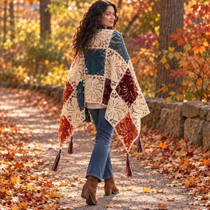 May include: A crochet shawl featuring a patchwork of square motifs in cream, dark teal, and burnt orange. The garment has a triangular shape with long, multi-colored tassels attached to the corners and edges, worn over blue denim jeans.