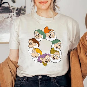 May include: A cream-coloured t-shirt with a circular graphic of the Seven Dwarfs from Snow White. Each dwarf has a distinct hat colour: green, orange, brown, yellow, purple, and green. The cartoon characters are smiling.