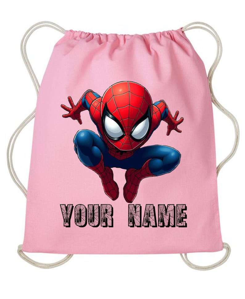 May include: A pink drawstring bag featuring a colorful illustration of Spider-Man in a crouching pose. The superhero is depicted in his classic red and blue suit. The text "YOUR NAME" is printed in black at the bottom.