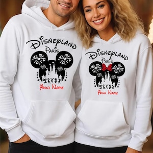 以下が含まれることがあります： 白いパーカー。ディズニーランドパリのグラフィックデザイン。ミッキーマウスの頭のシルエットに花火、お城、そして「Disneyland Paris」と「Your Name」の文字が黒と赤で描かれています。1つのパーカーには赤いリボンが付いています。