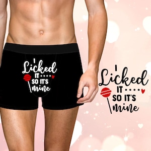 May include: Black boxer shorts with the text "Licked it... so it's mine" in white script. A red lollipop graphic is included. The waistband is black.