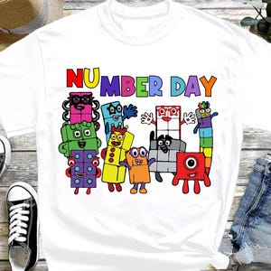 May include: White t-shirt featuring the text "NUMBER DAY" in colorful letters above a group of cartoon number characters. The characters are various colors and shapes, with smiling faces and arms.