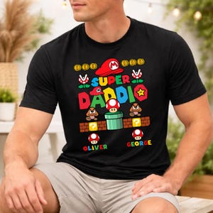 May include: Black t-shirt featuring a colorful Super Mario-themed design with the text "SUPER DADDI". The design includes Mario's hat, coins, and characters. The names "OLIVER" and "GEORGE" are also printed on the shirt.