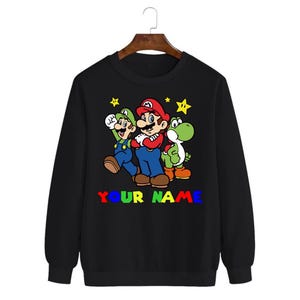 Peut inclure: Sweat-shirt noir à col rond avec un graphisme coloré de Mario, Luigi et Yoshi. Le texte "YOUR NAME" est imprimé en couleurs arc-en-ciel sous les personnages. Des étoiles jaunes sont en arrière-plan.