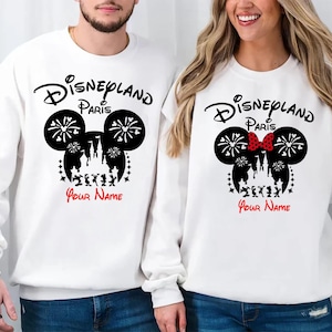 Op de afbeelding: Witte sweatshirts met een Disneyland Paris-ontwerp. Het ontwerp toont een zwarte Mickey Mouse-kop met vuurwerk, een kasteelsilhouet en de tekst "Disneyland Paris" erboven en "Your Name" in het rood eronder. Eén sweatshirt heeft een rode strik.