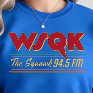 Może przedstawiać: Niebieska bluza z napisem "WSQK The Squawk 94.5 FM" w kolorze czerwonym, żółtym i pomarańczowym. Projekt zawiera błyskawicę i poziome linie w różnych kolorach. Widoczny jest srebrny naszyjnik.