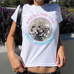 Può includere: T-shirt bianca con grafica a sfera da discoteca argentata e la scritta "KISS ALL THE TIME. DISCO, OCCASIONALLY." La sfera da discoteca è composta da piccoli quadrati riflettenti. Il testo è in rosa e blu.